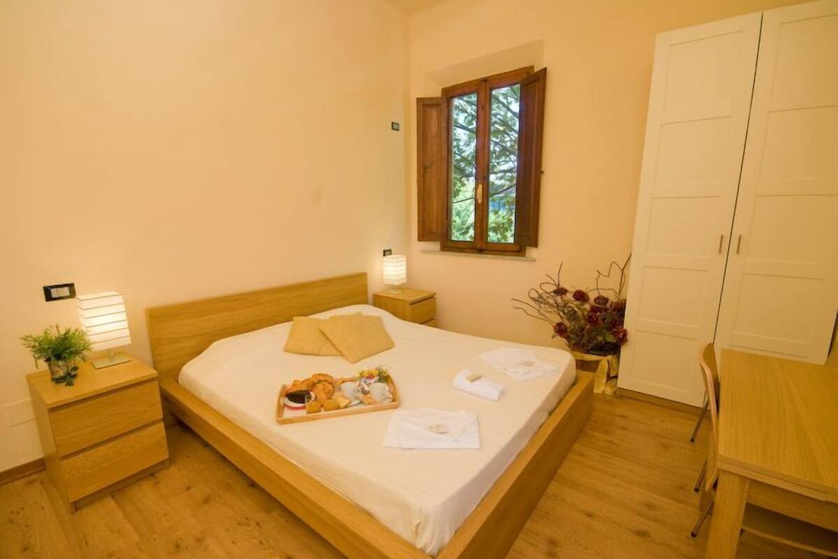 A neatly made bed with a tray of breakfast items, a bedside lamp, and a window with a view of trees.