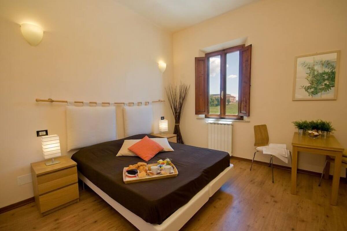 A bedroom with a bed, bedside tables, a window with curtains, a desk with a chair, and a small table with plants.