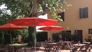 An outdoor patio with multiple wooden tables and chairs, red umbrellas, and a tree providing shade. - B&B Dolce Notte (Pisa)