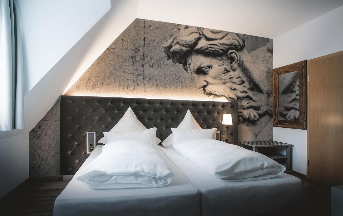 Une chambre d’hôtel moderne avec un grand lit, une tête de lit capitonnée, une œuvre d’art accrochée au mur et une table de chevet avec une lampe.