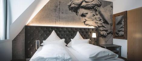 Una habitación de hotel moderna con una cama grande, un cabecero capitonado, una obra de arte colgada en la pared y una mesita de noche con una lámpara.