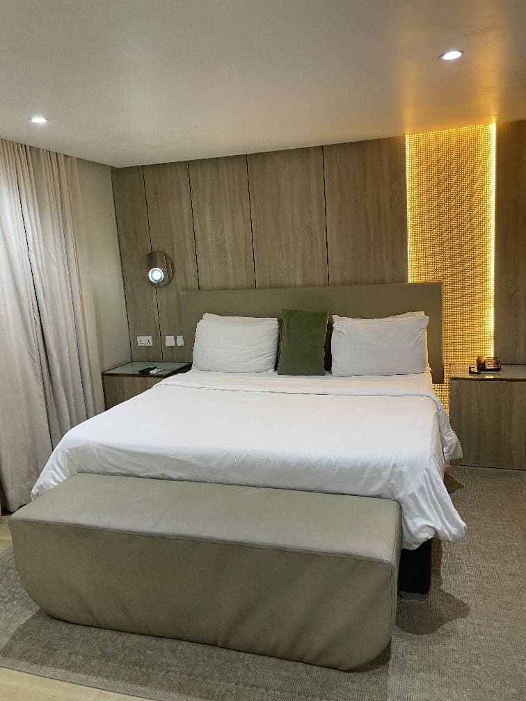 A hotel room with a large bed, a bedside table, a nightstand, a bench, and a wall-mounted light fixture.