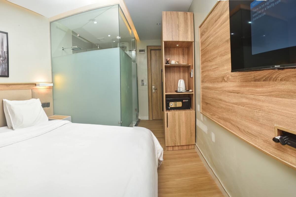 A modern hotel room with a large bed, bedside lamps, a flat-screen TV, a glass shower enclosure, and a wooden wardrobe.