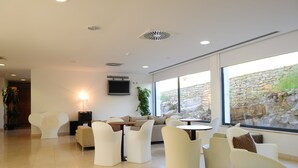 A modern lounge area with white furniture, a flat-screen TV, and large windows offering a view of a stone wall and greenery. - HI Alvados - Pousada de Juventude - Hostel (Porto de Mos)