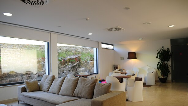 A modern living room with a beige sofa, white chairs, a stone wall view, and a potted plant. - HI Alvados - Pousada de Juventude - Hostel (Porto de Mos)
