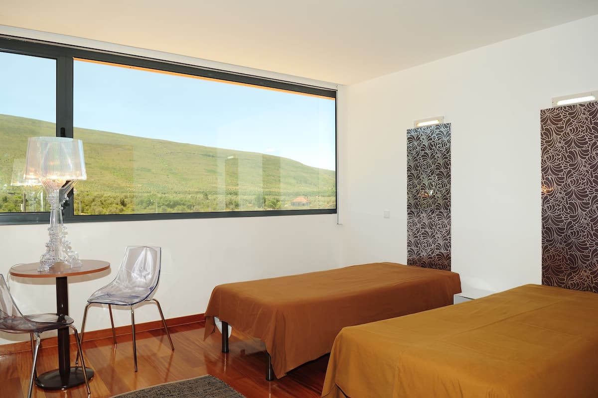 A hotel room with two beds, a table with chairs, a lamp, and a large window offering a view of a green landscape.