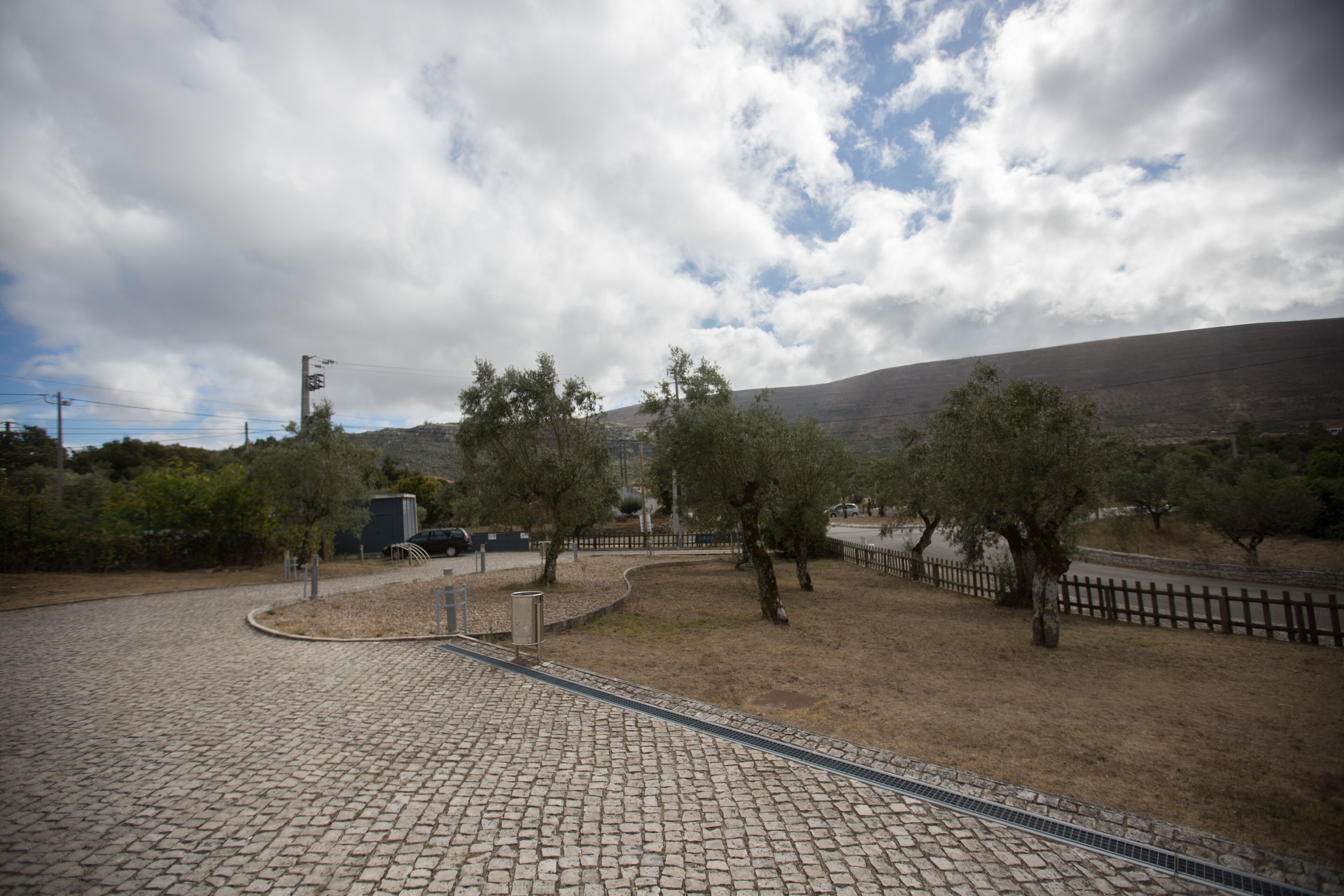 Ein gepflasterter Weg führt zu einem von Zäunen umschlossenen Bereich mit Bäumen und einem entfernten Berg.