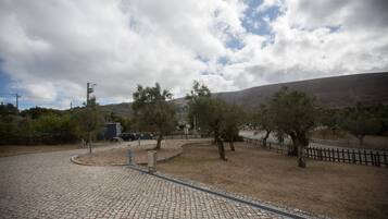 Ein gepflasterter Weg führt zu einem von Zäunen umschlossenen Bereich mit Bäumen und einem entfernten Berg.