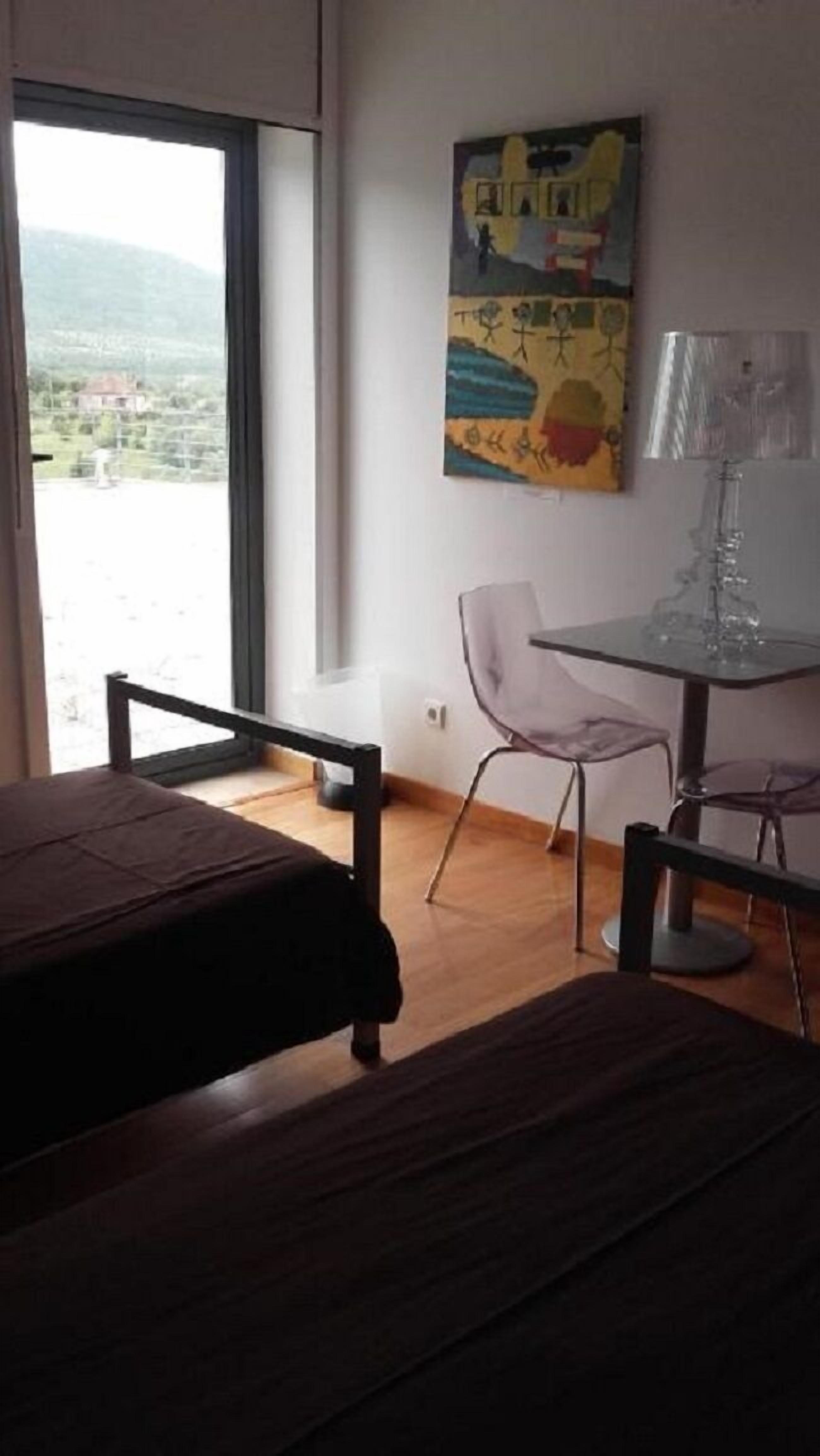 Una habitación con cama, una mesa y una silla. Se ve una ventana con vistas a una casa y montañas.