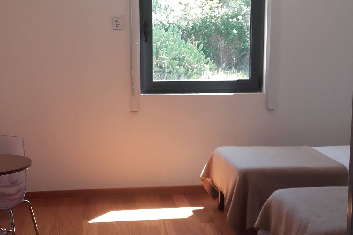 A room with a large window, wooden flooring, two beds, a chair, and a small table.