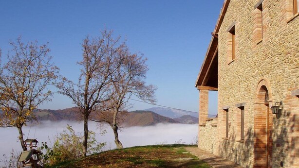 A stone building with a stone roof, a small garden with a bench, and a view of a mountain range through a misty valley. - Girfalco - Country House - Bed&Breakfast (Urbino)