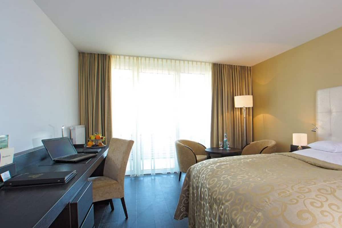 Habitación de hotel con cama, escritorio con computadora portátil, silla, mesita de noche con lámpara y vistas al exterior a través de amplias ventanas.