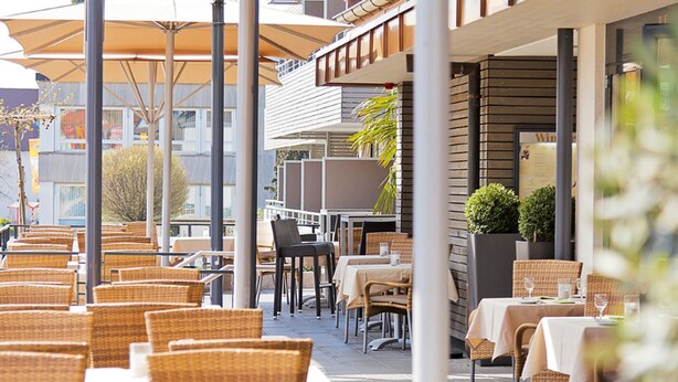 An outdoor cafe with wicker chairs and tables, umbrellas, and a building with a balcony in the background.