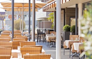 Ein Straßencafé mit Korbstühlen und Tischen, Sonnenschirmen und einem Gebäude mit Balkon im Hintergrund.