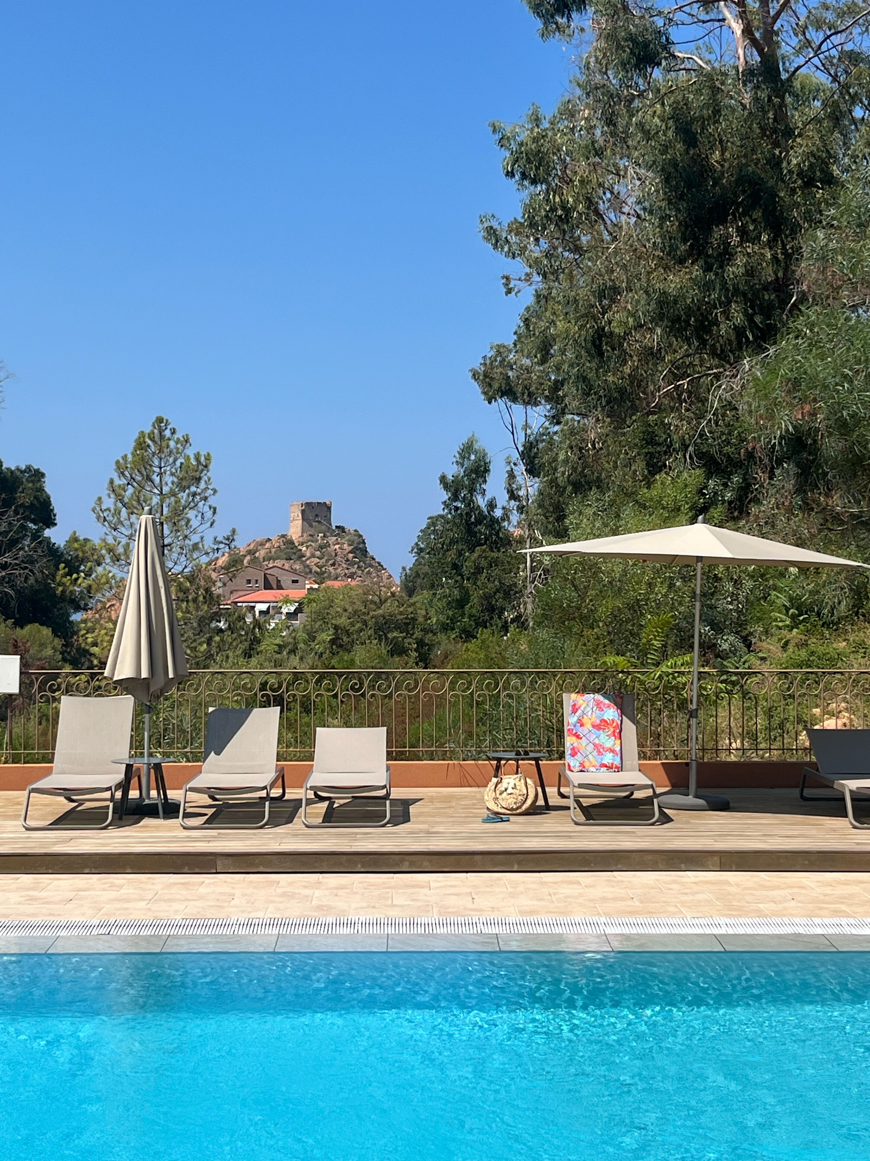 Un espace aménagé au bord de la piscine, avec des chaises longues, des parasols et une vue sur une colline où se dresse une structure ressemblant à un château.