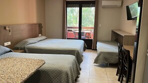 Habitación de hotel con tres camas, un escritorio, un televisor y un balcón con mobiliario exterior.