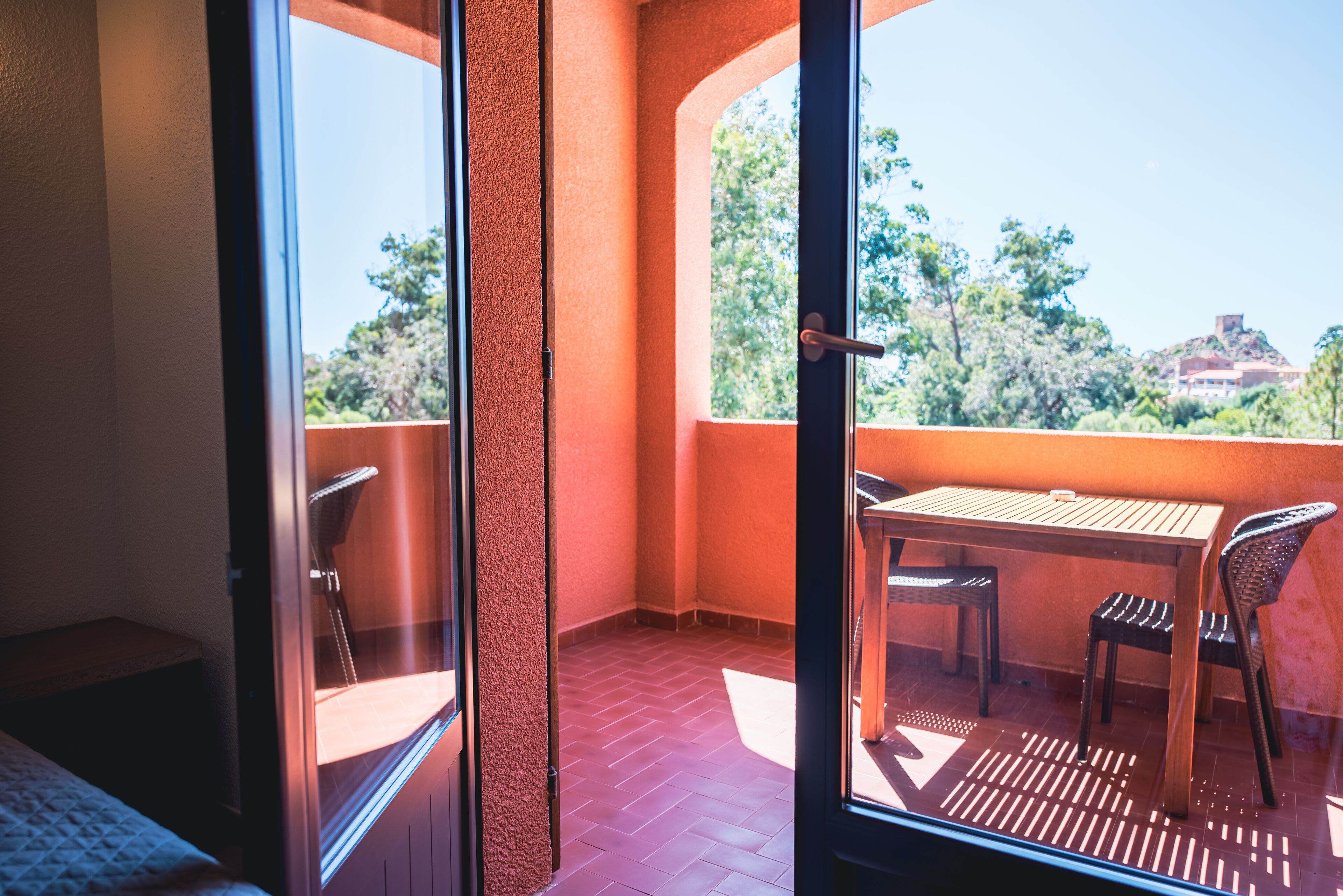 Uma varanda com mesa e cadeiras, uma porta que dá para um quarto e vista para árvores e uma colina.