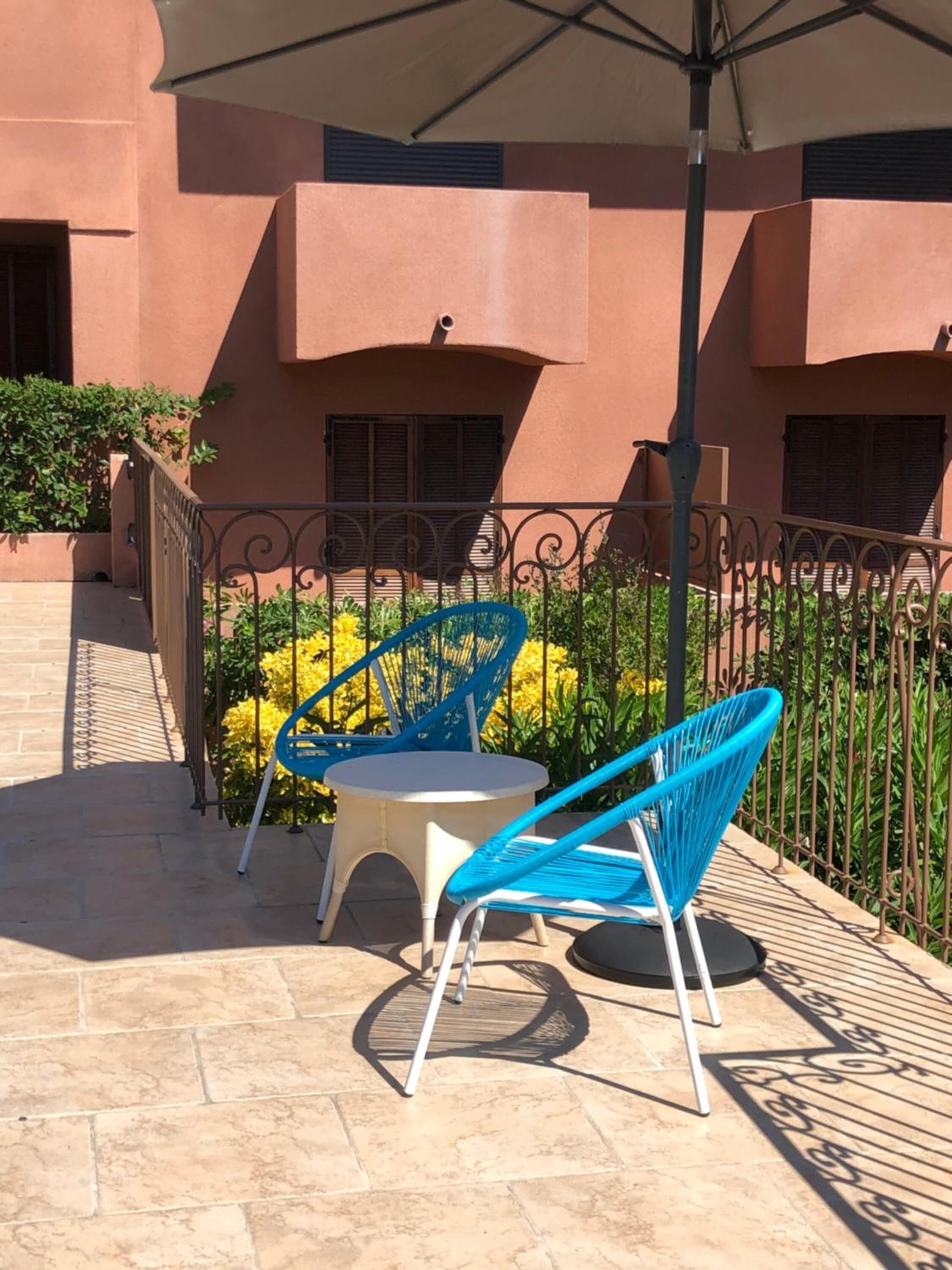 Un patio exterior con dos sillas azules y una pequeña mesa redonda, una cerca de metal y un edificio con fachada marrón rojiza.