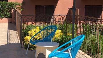 Un patio exterior con dos sillas azules y una pequeña mesa redonda, una cerca de metal y un edificio con fachada marrón rojiza.