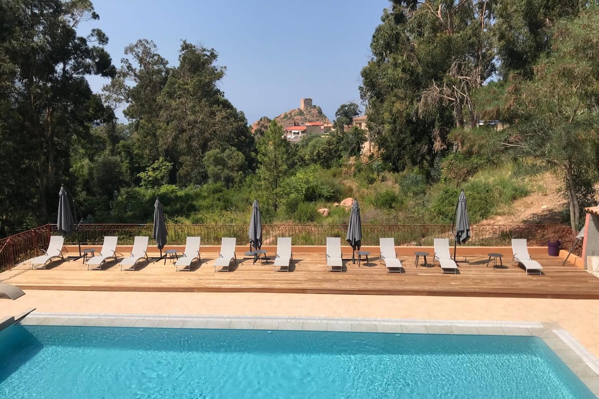 A poolside area with lounge chairs and umbrellas, surrounded by trees and a hill with a building in the background.