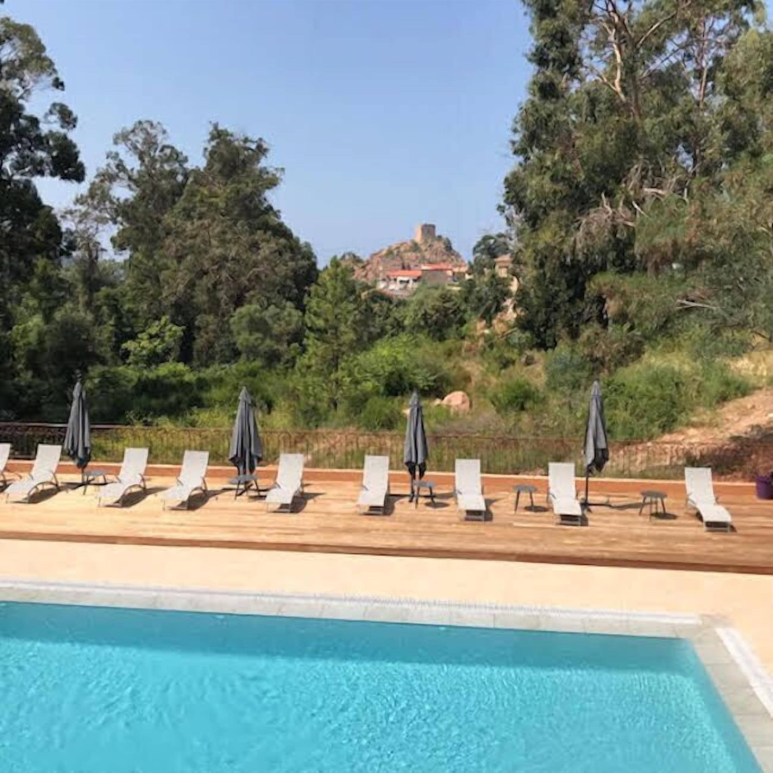 A pool with lounge chairs and umbrellas, surrounded by trees and a view of a hill with buildings.