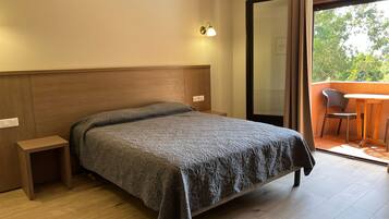 Habitación de hotel con cama, mesita de noche, lámpara de pared y vistas al exterior a través de un ventanal.
