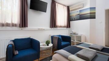 Una habitación de hotel moderna con una cama, dos sillones azules, una mesita redonda con una planta, un televisor montado en la pared y una ventana con cortinas.
