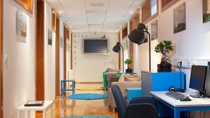 A modern hotel room with a wooden floor, a desk with a computer, a blue chair, a sofa, and a TV mounted on the wall. - Hostel Bureau (Zagreb)