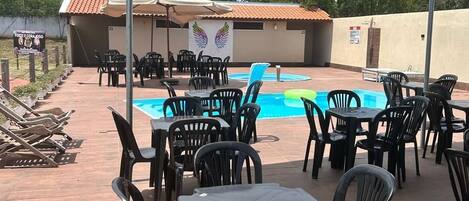 Un patio al aire libre con piscina, rodeado de sillas y mesas de plástico, bajo una zona cubierta.