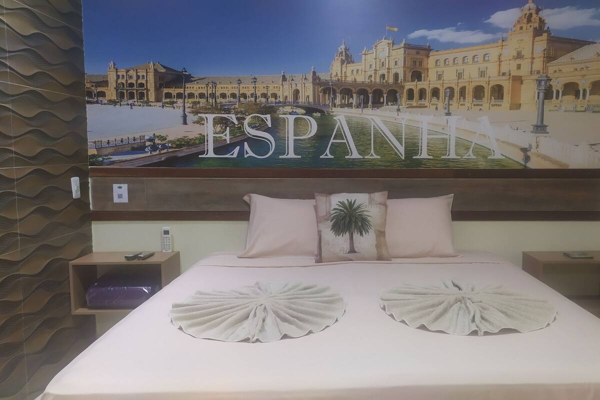 Une chambre d’hôtel avec un grand lit, deux tables de chevet, un téléphone fixé au mur et une affiche représentant un bâtiment sur lequel on peut lire le mot « ESPAÑI ».