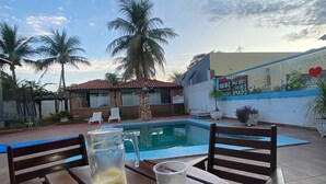 Mesa junto a la piscina con una jarra de jugo de naranja y un vaso, un plato con tostadas, y una vista de una alberca con palmeras y un edificio con un mural al fondo.