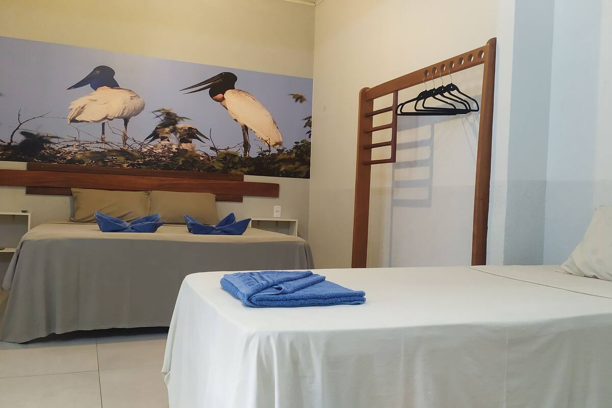 Une chambre d’hôtel avec deux lits, une tête de lit en bois, un porte-manteau fixé au mur et un tableau représentant des oiseaux.