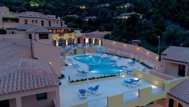 A resort with a swimming pool surrounded by lounge chairs and umbrellas, set against a backdrop of terraced buildings and lush greenery. - Sosventos Village (Teulada)