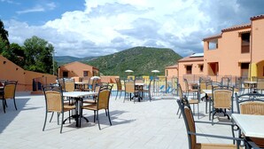 An outdoor patio with tables and chairs, a swimming pool, and buildings in the background. - Sosventos Village (Teulada)