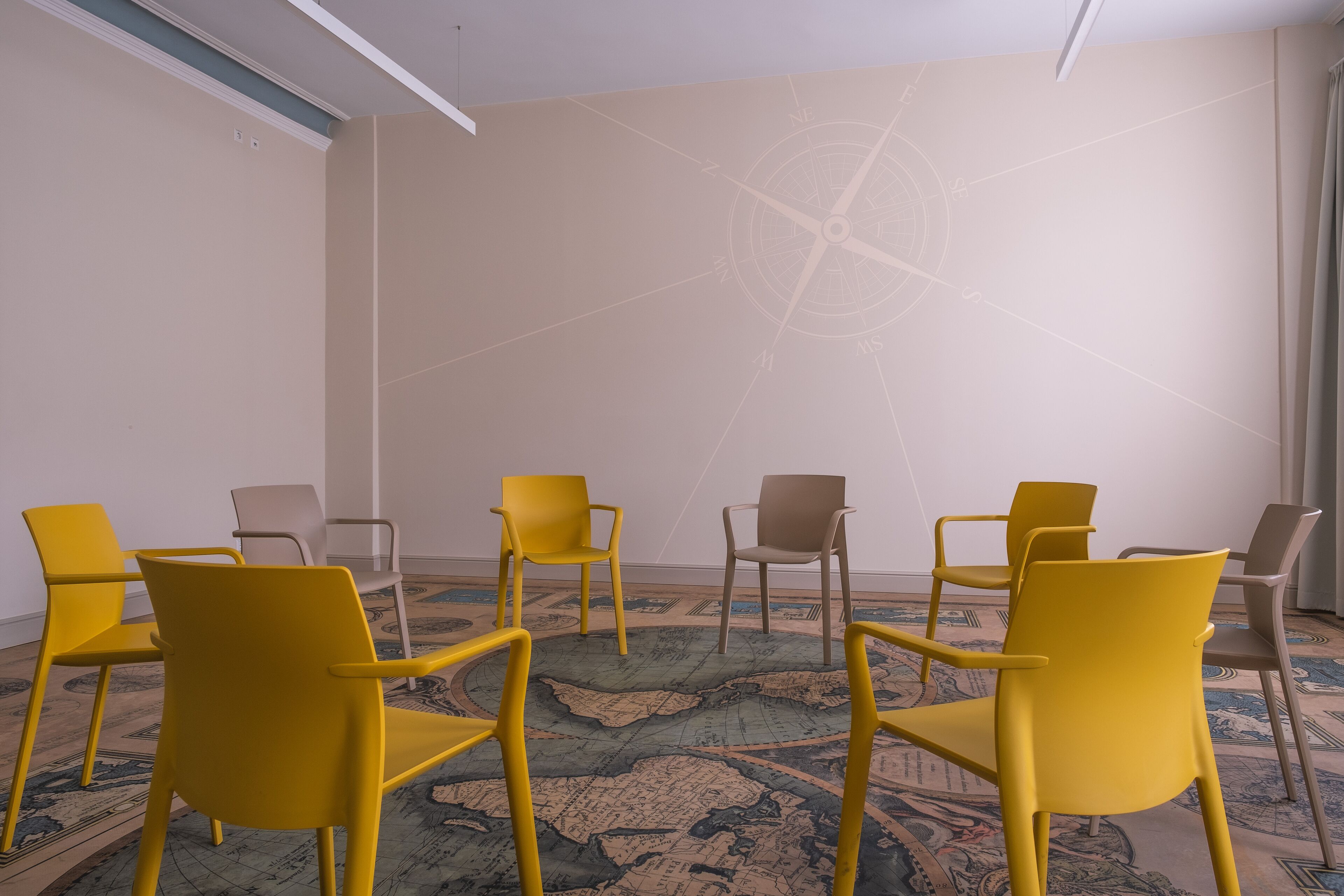 Een vergaderruimte met een vloerbedekking, een kompas als wanddecoratie, en gele en beige stoelen die in een halve cirkel zijn opgesteld.
