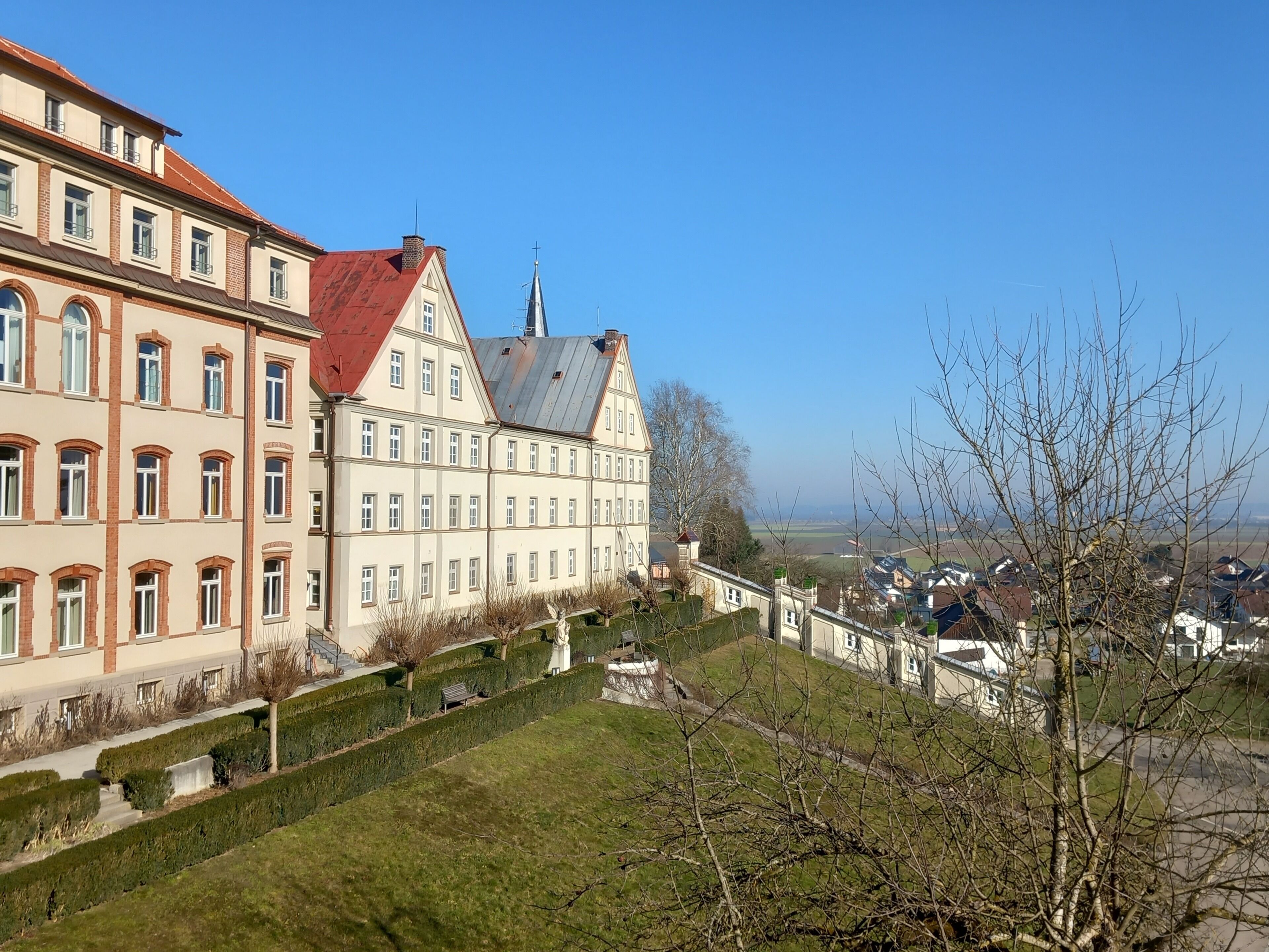 A large, historic building with multiple floors and a red roof, surrounded by well-maintained gardens and a clear view of the landscape.