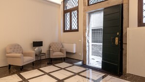 Un'ampia hall con pavimento a scacchi, due poltrone, un tavolino con una lampada e un'imponente porta.