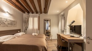 Una camera d'albergo con soffitto in legno, un letto con coperta color beige, una scrivania con lampada e una sedia.