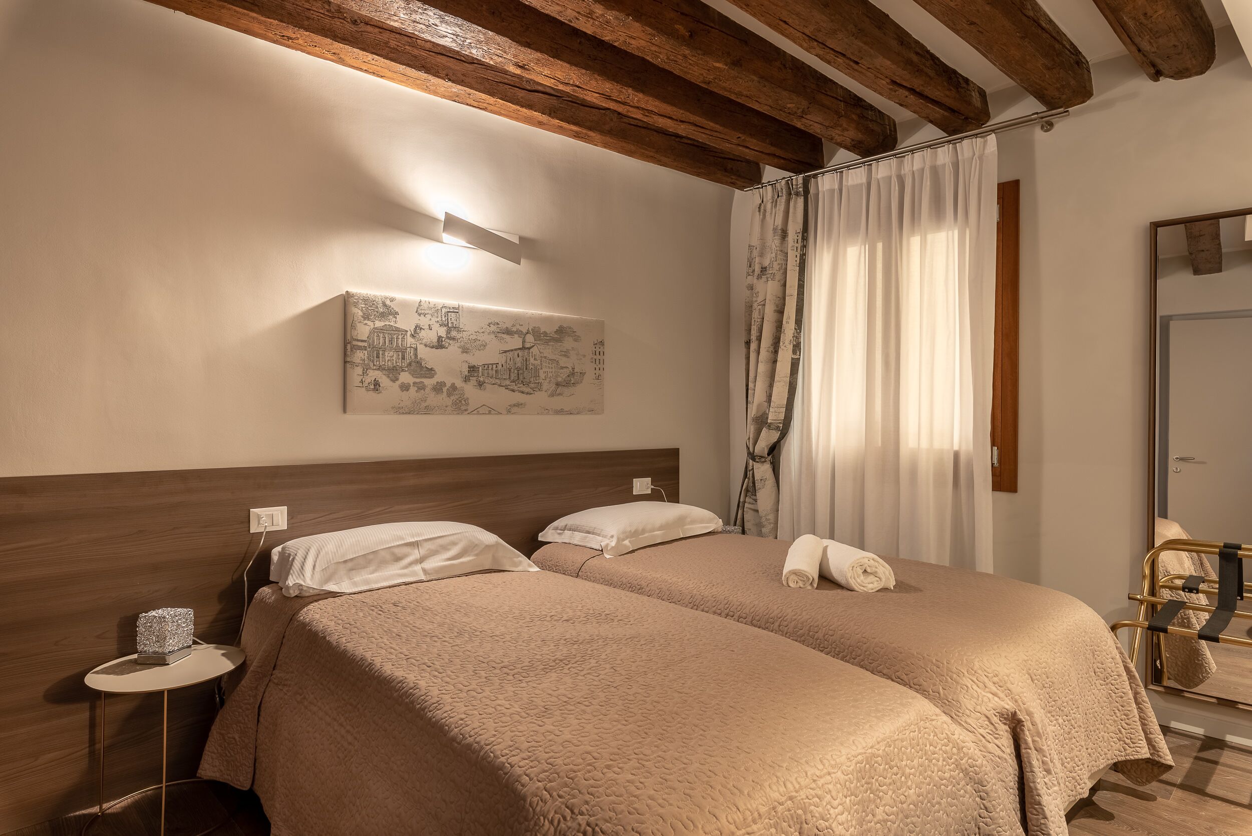 Quarto de hotel com duas camas, vigas de madeira, uma mesa pequena com abajur e um quadro na parede.