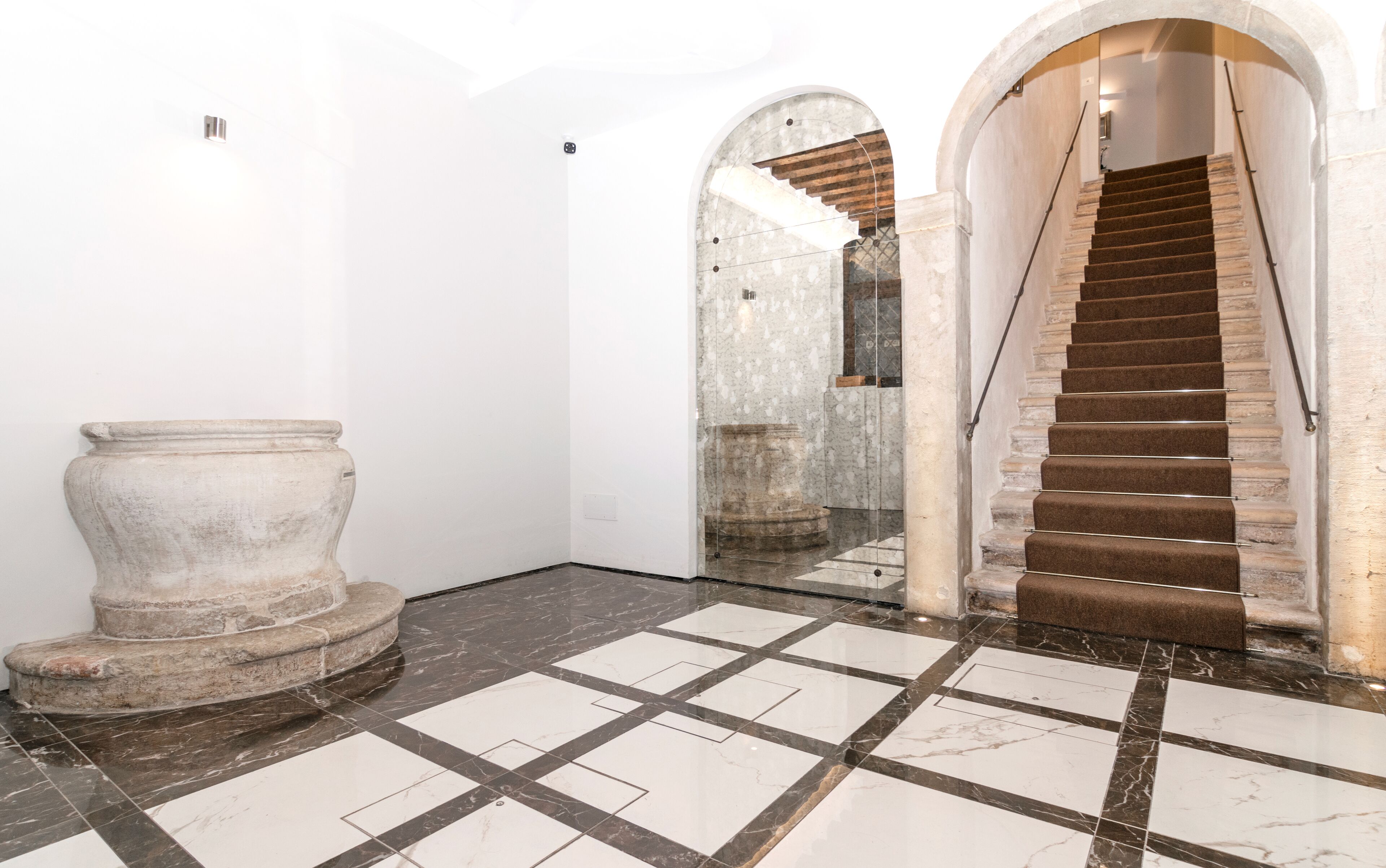 Um corredor com piso de mármore, um pedestal de pedra e uma escada com carpete marrom.