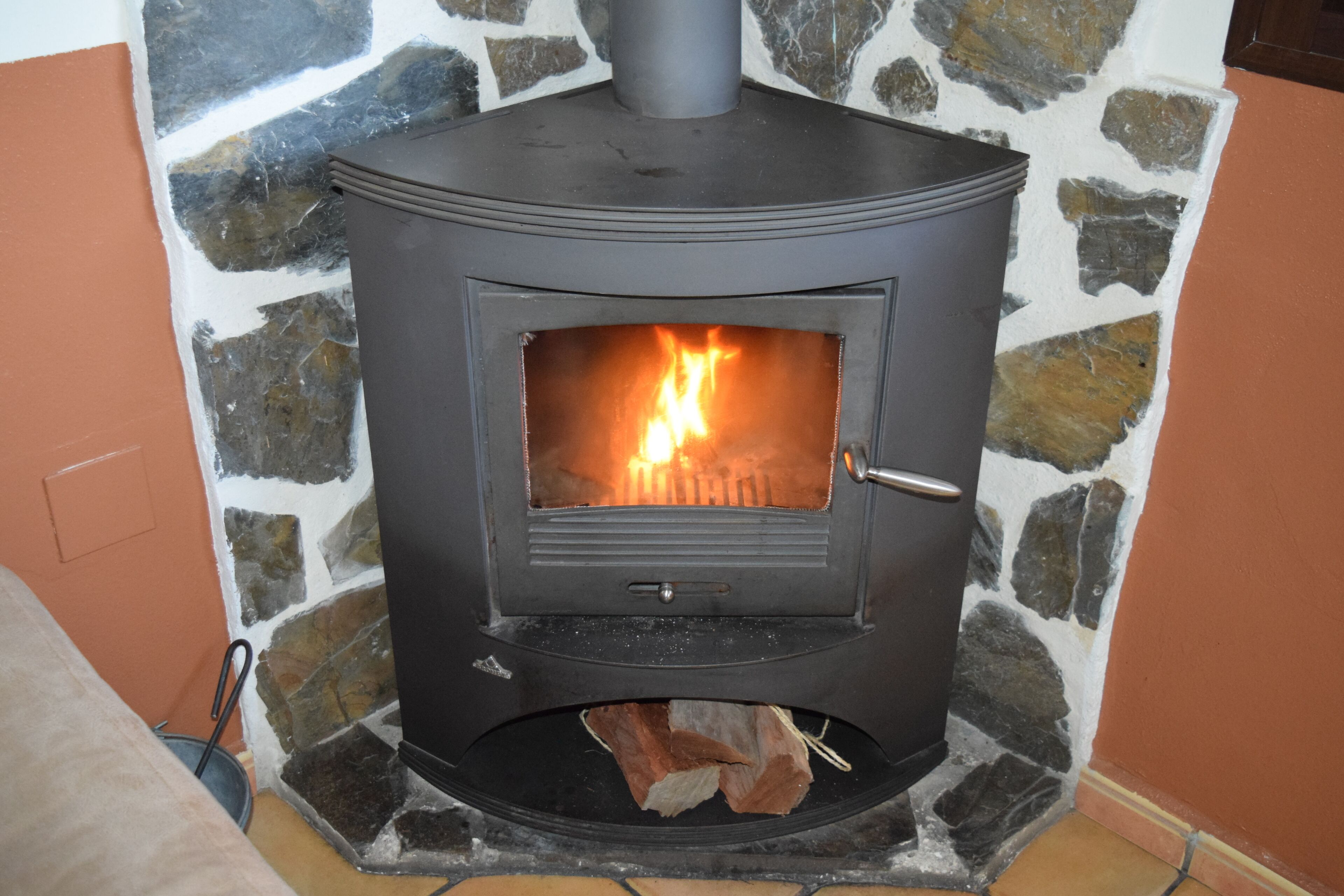 A wood-burning stove with a stone wall backdrop, a door handle, and a visible fire inside.