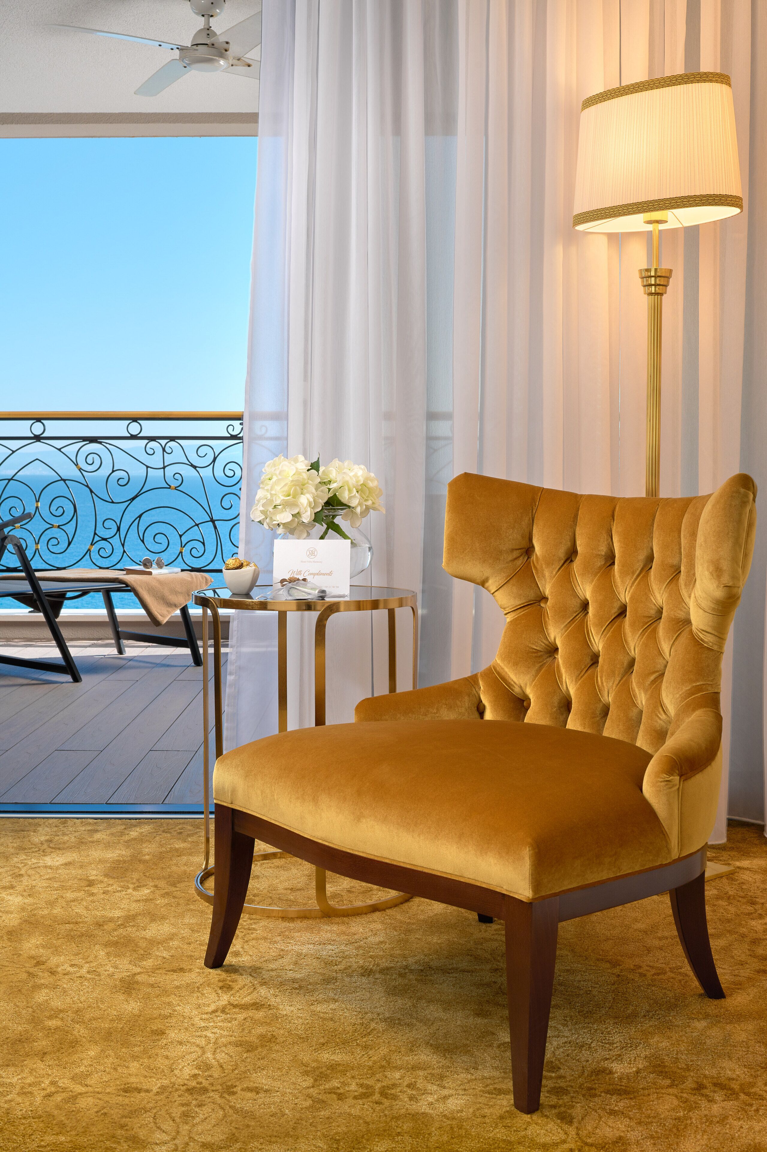 A plush, tufted armchair in a room with a view of the ocean, a standing lamp, and a small table with a vase of flowers.