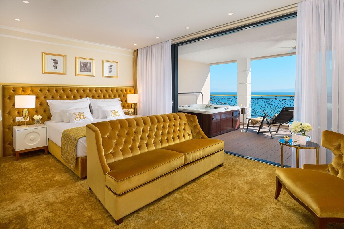 A hotel room with a bed, a sofa, a desk, and a balcony with a view of the sea.