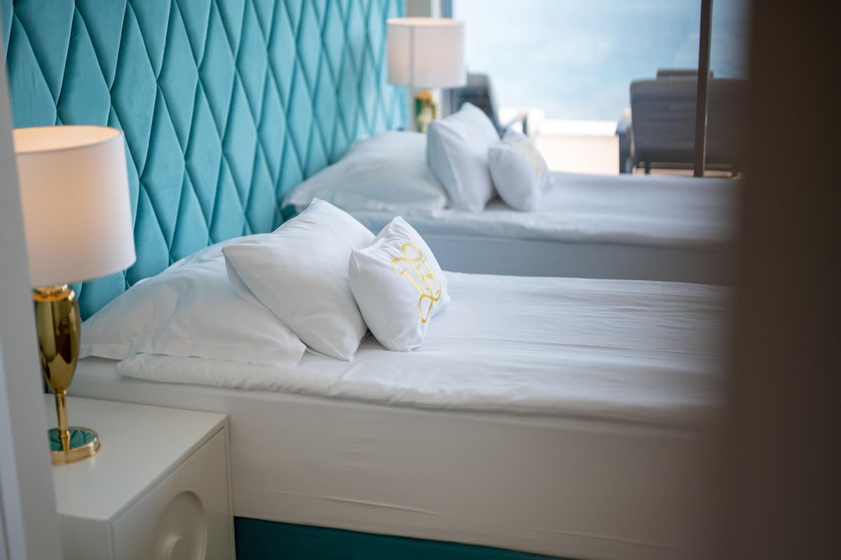 A hotel room with two beds, a teal headboard, white bedding, and a gold lamp on the nightstand.