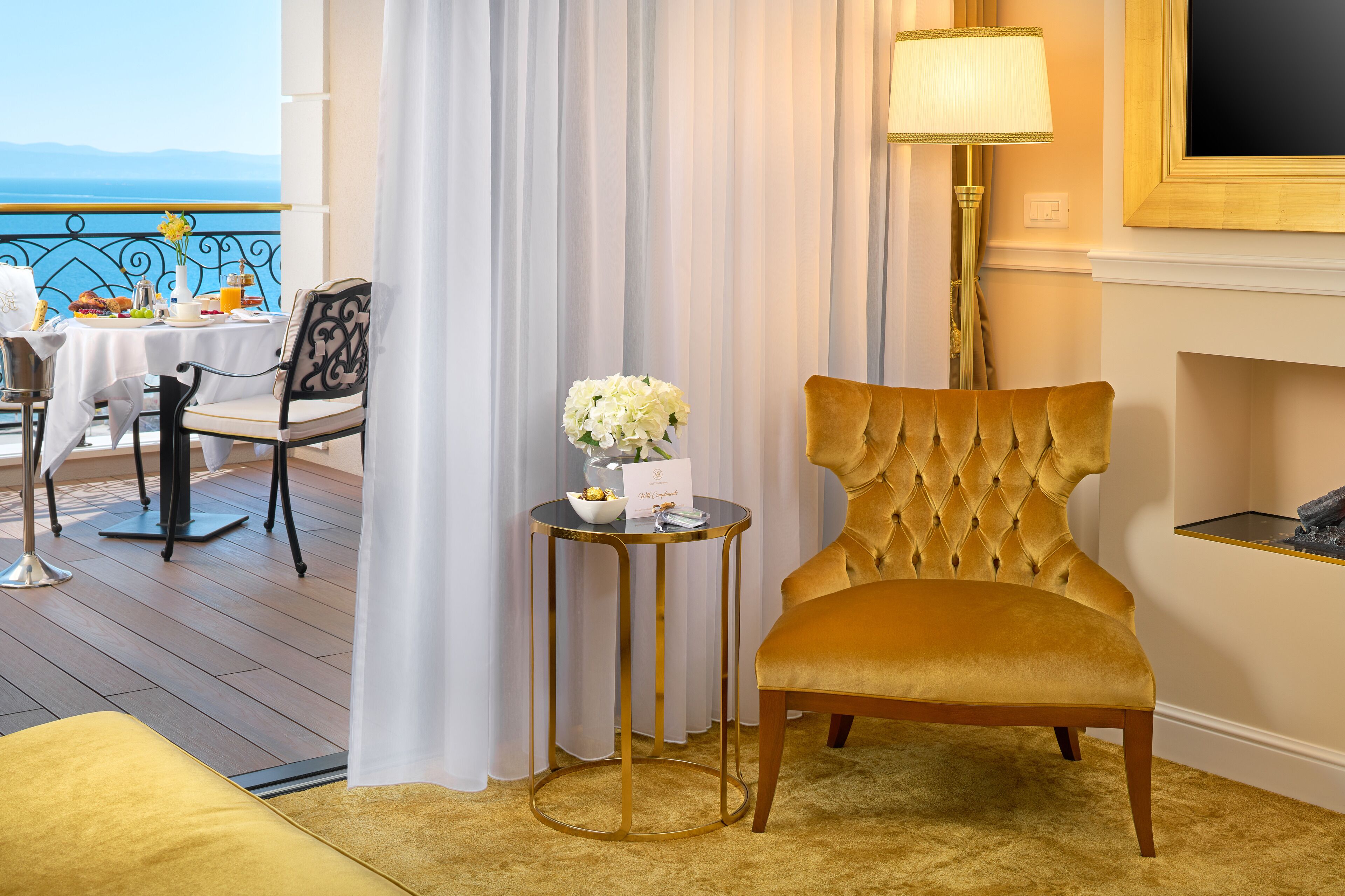 Een hotelkamer met uitzicht op zee, een eethoek met een gedekte tafel voor twee personen, een goudkleurige fauteuil en een bijzettafeltje met een bloevas.