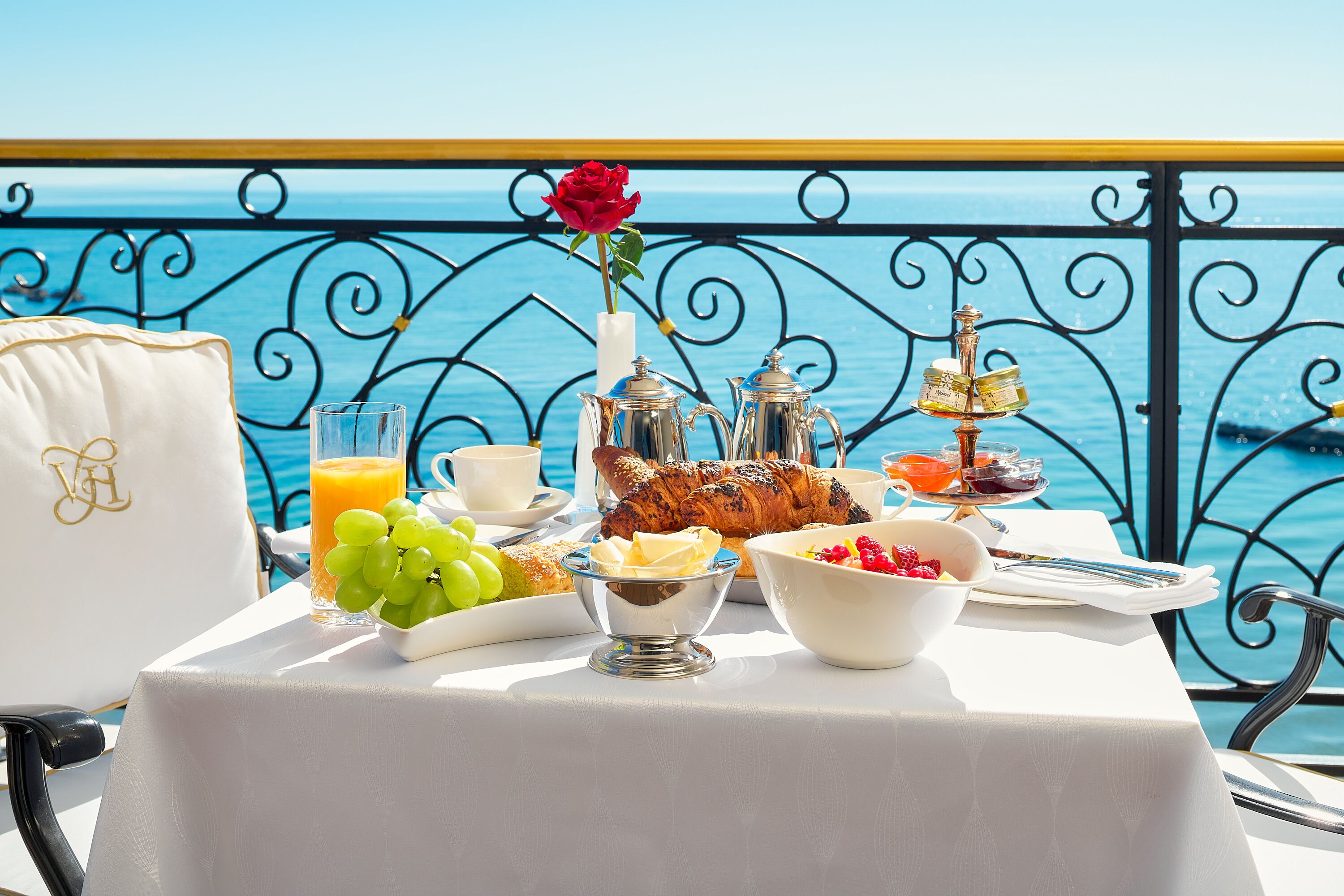 Une table de petit-déjeuner garnie de divers aliments, un verre de jus d’orange et une rose dans un vase, le tout mis en valeur par une vue panoramique sur l’océan.