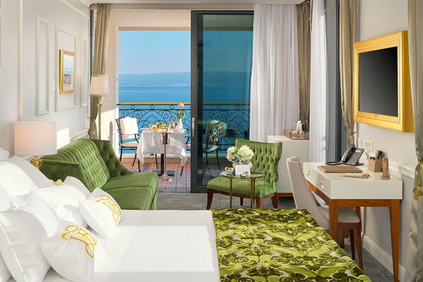 A hotel room with a green sofa, a white bed, a desk with a phone, and a view of the sea. - Hotel Villa Harmony (Split)