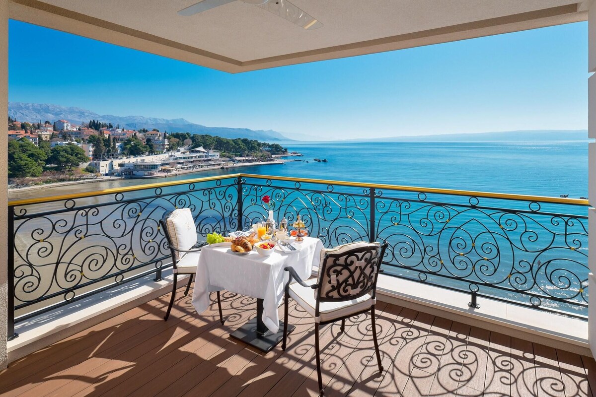 A balcony with a table set for two, overlooking a coastal view with mountains in the distance.
