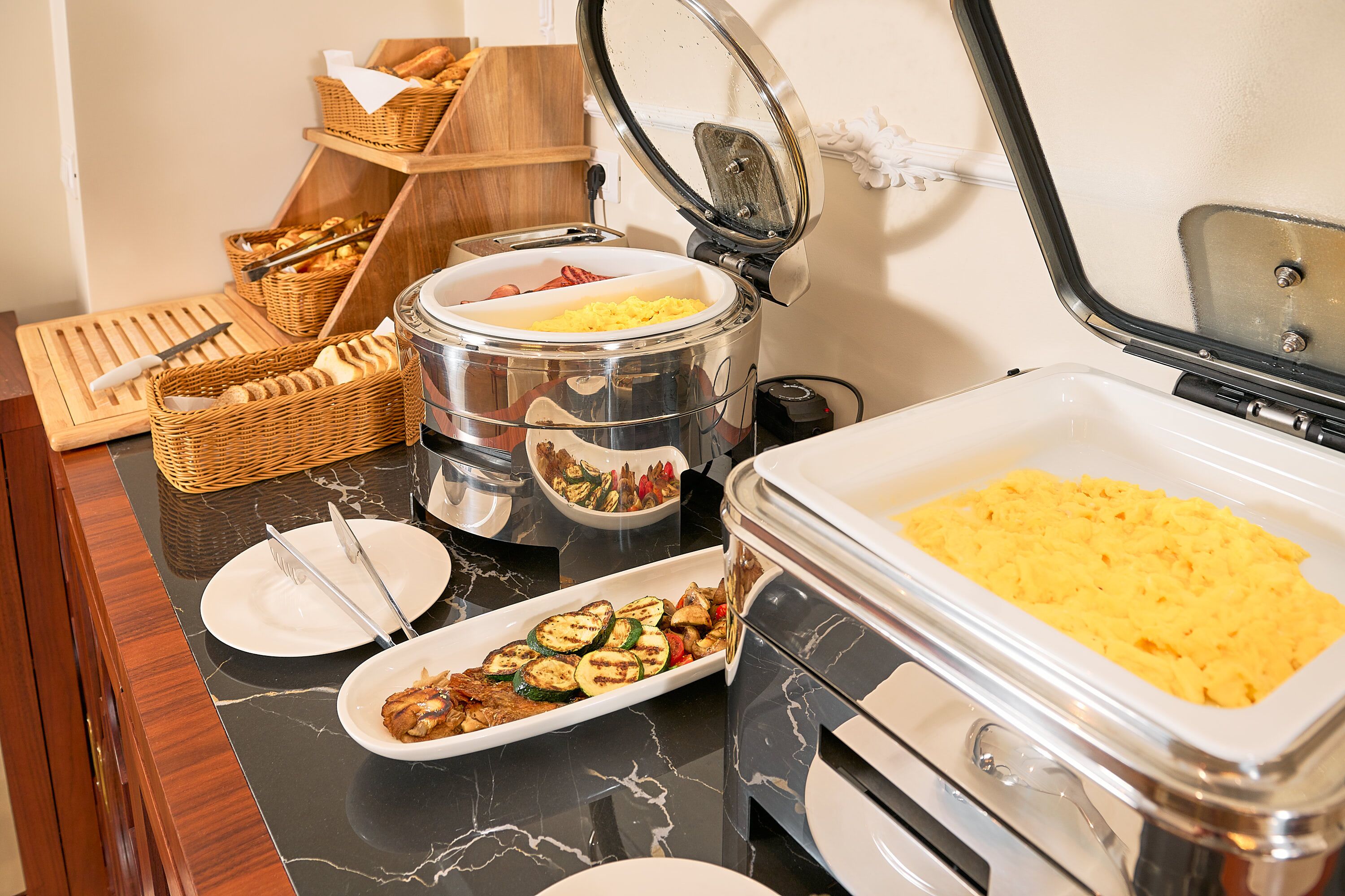 A buffet setup with various food items, including scrambled eggs, grilled vegetables, and a meat dish, served on a marble countertop.