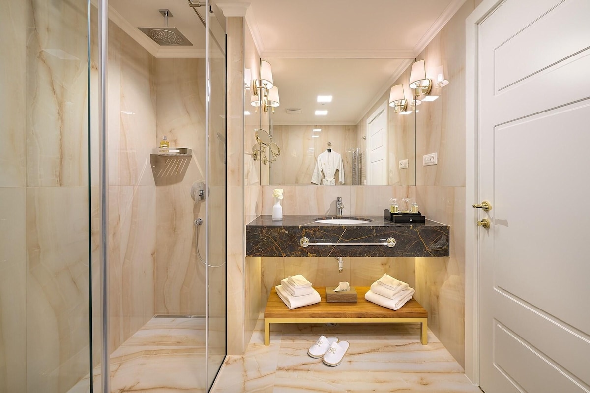 A modern bathroom with a glass-enclosed shower, a vanity with a marble countertop, a wooden bench with towels, and a large mirror.